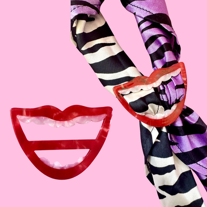 A fashion accessory set featuring a red translucent lip-shaped hair clip and a matching red lip-shaped comb, displayed alongside a purple, black, and white zebra-print silk scarf on a solid pink background.