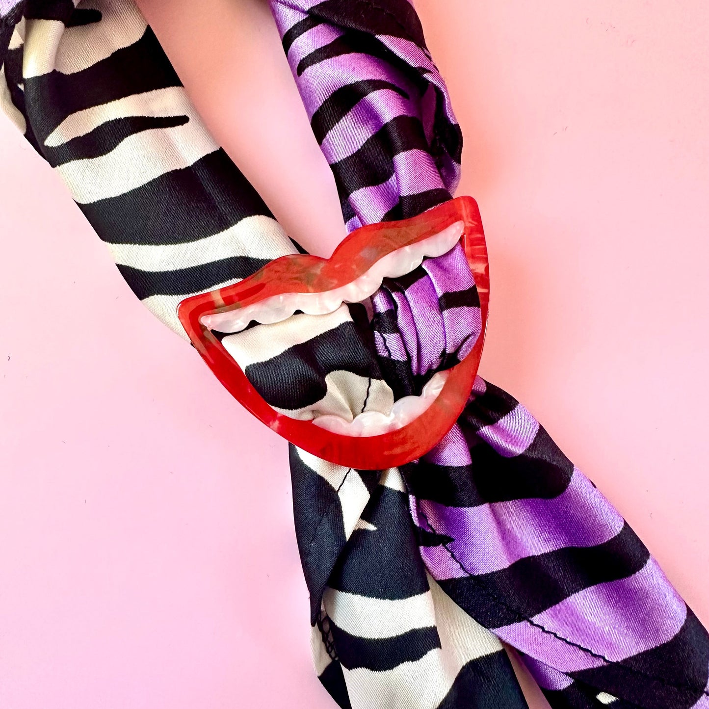 A close-up of a scarf tied in a knot with a red lip-shaped clip, featuring a black and white zebra print on one side and purple and black stripes on the other, set against a solid pink background.