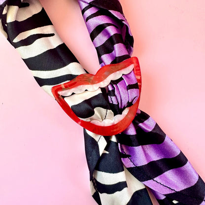 A close-up of a scarf tied in a knot with a red lip-shaped clip, featuring a black and white zebra print on one side and purple and black stripes on the other, set against a solid pink background.