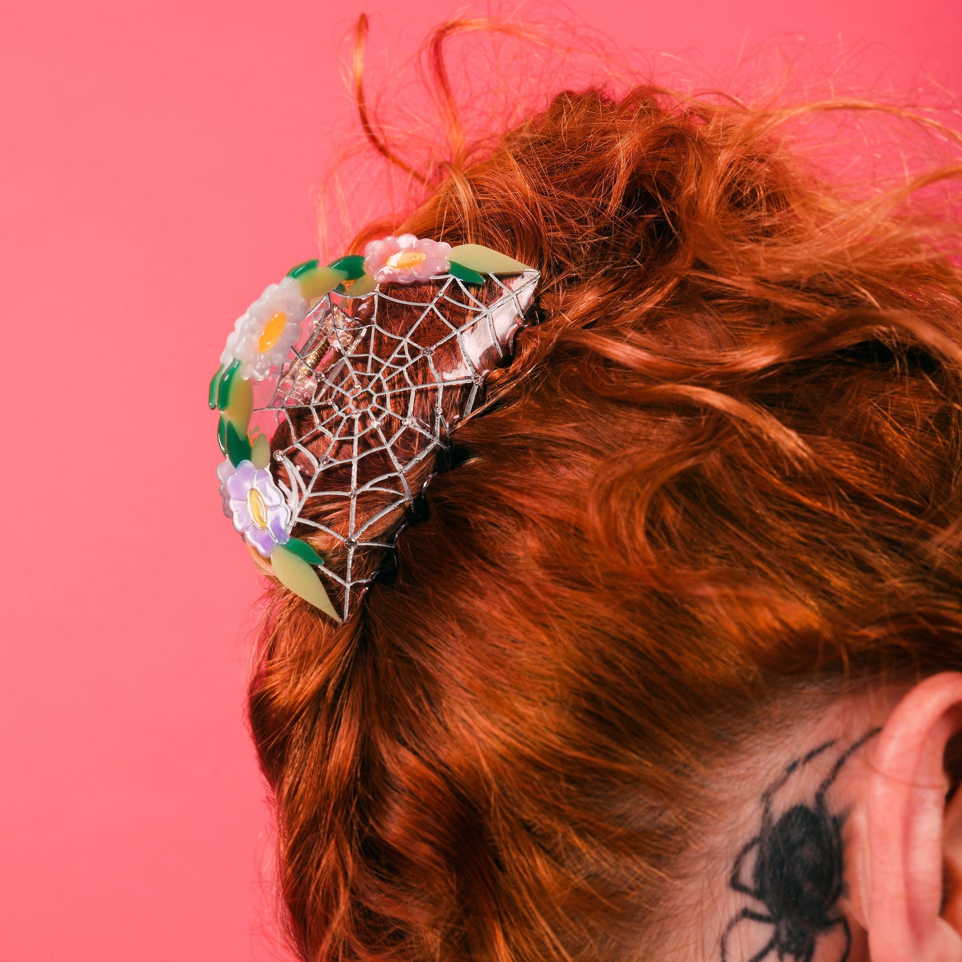 Woman with red hair and tattoos against a pink background wearing a clear spiderweb and rose floral hair claw clip as an updo with red hair.
