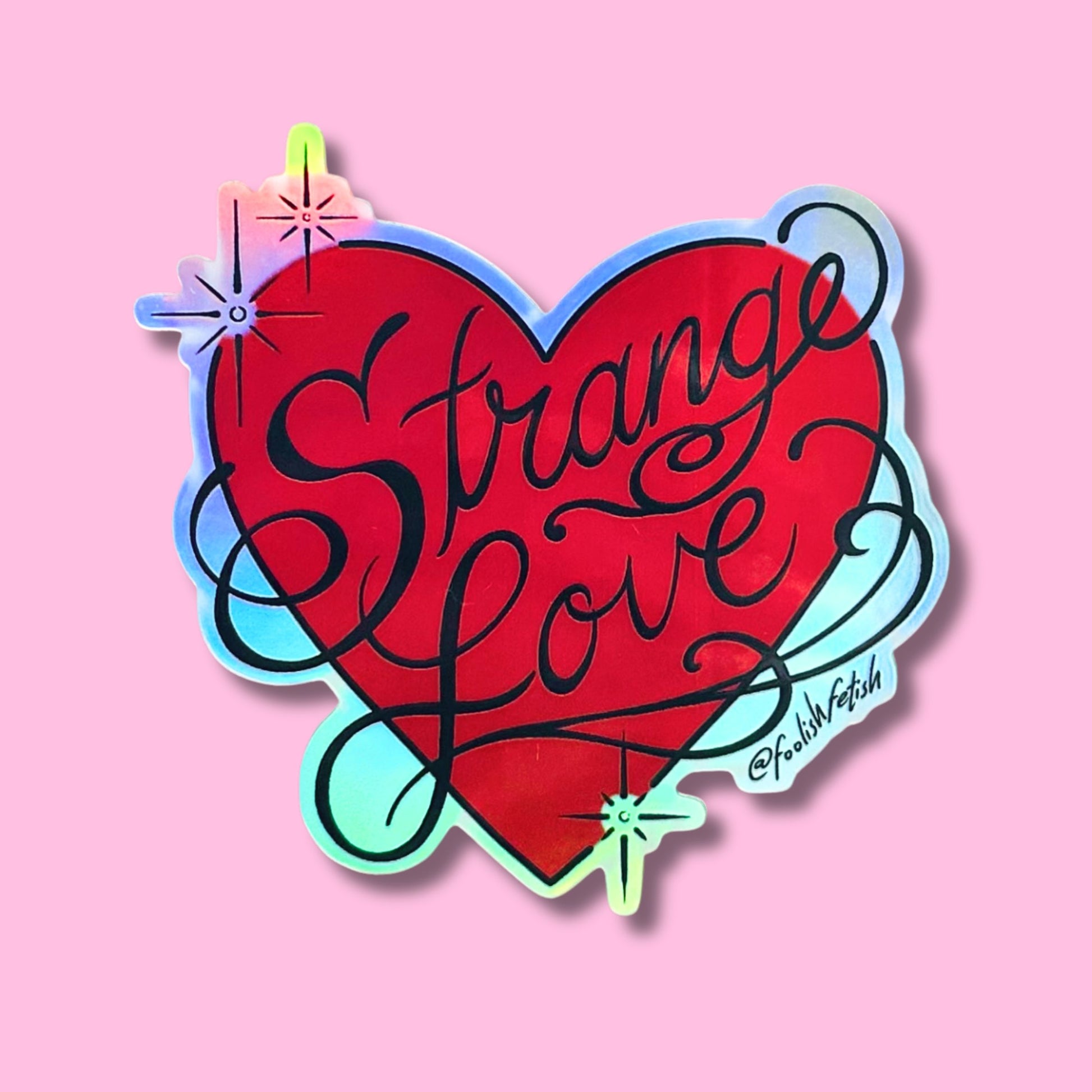 A die-cut sticker featuring a red heart with the words 'Strange Love' in black cursive script, outlined in iridescent pastel colors, shown on a solid pink background.