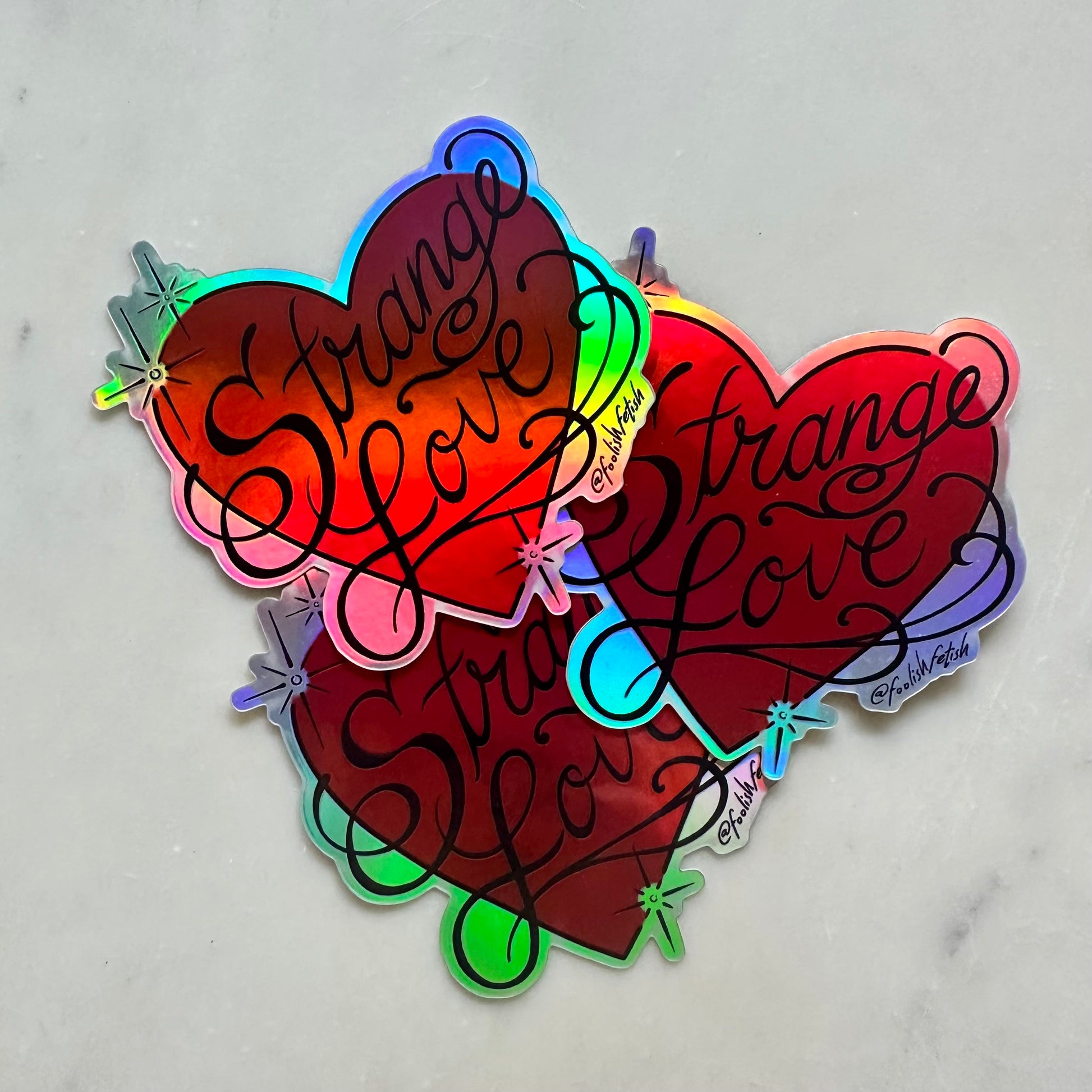 Three holographic heart-shaped stickers with the phrase 'strange love' in black cursive script, arranged on a light gray marble surface.