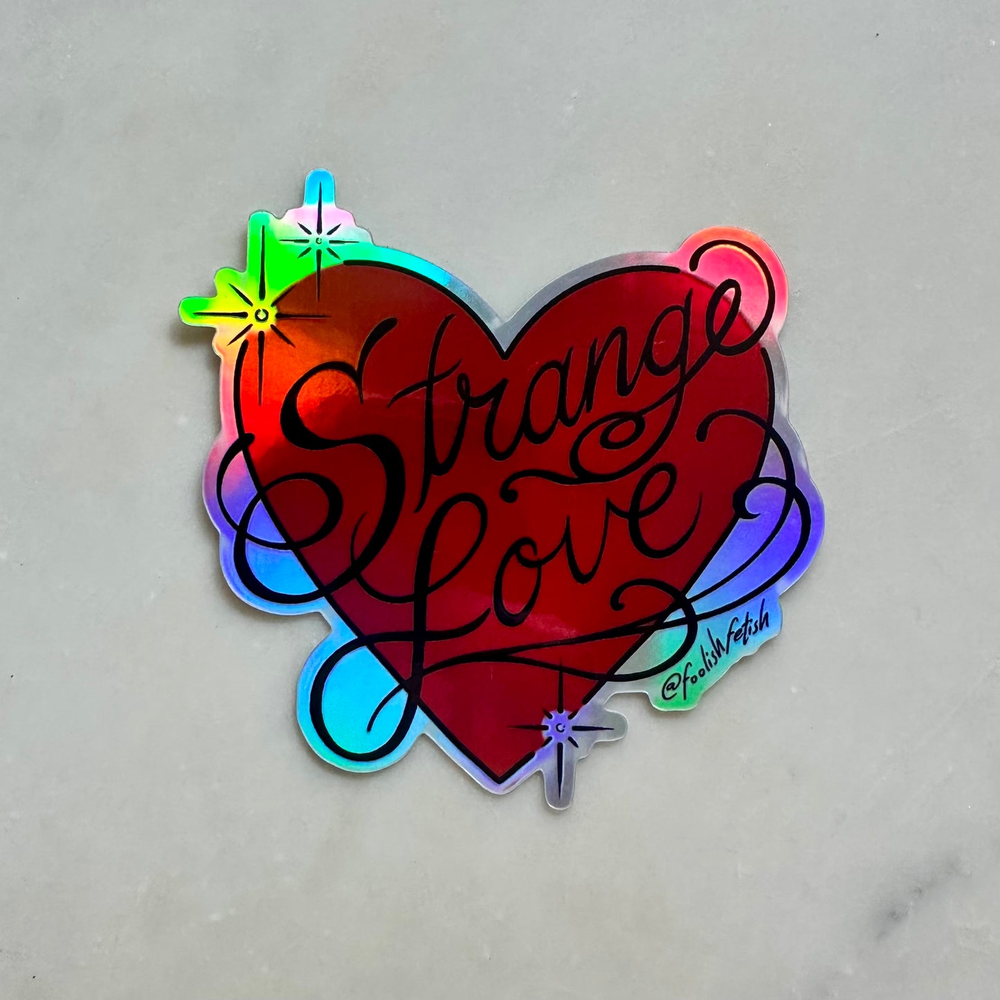 A holographic heart-shaped sticker with the words 'Strange Love' in black script, placed on a light gray marble surface.