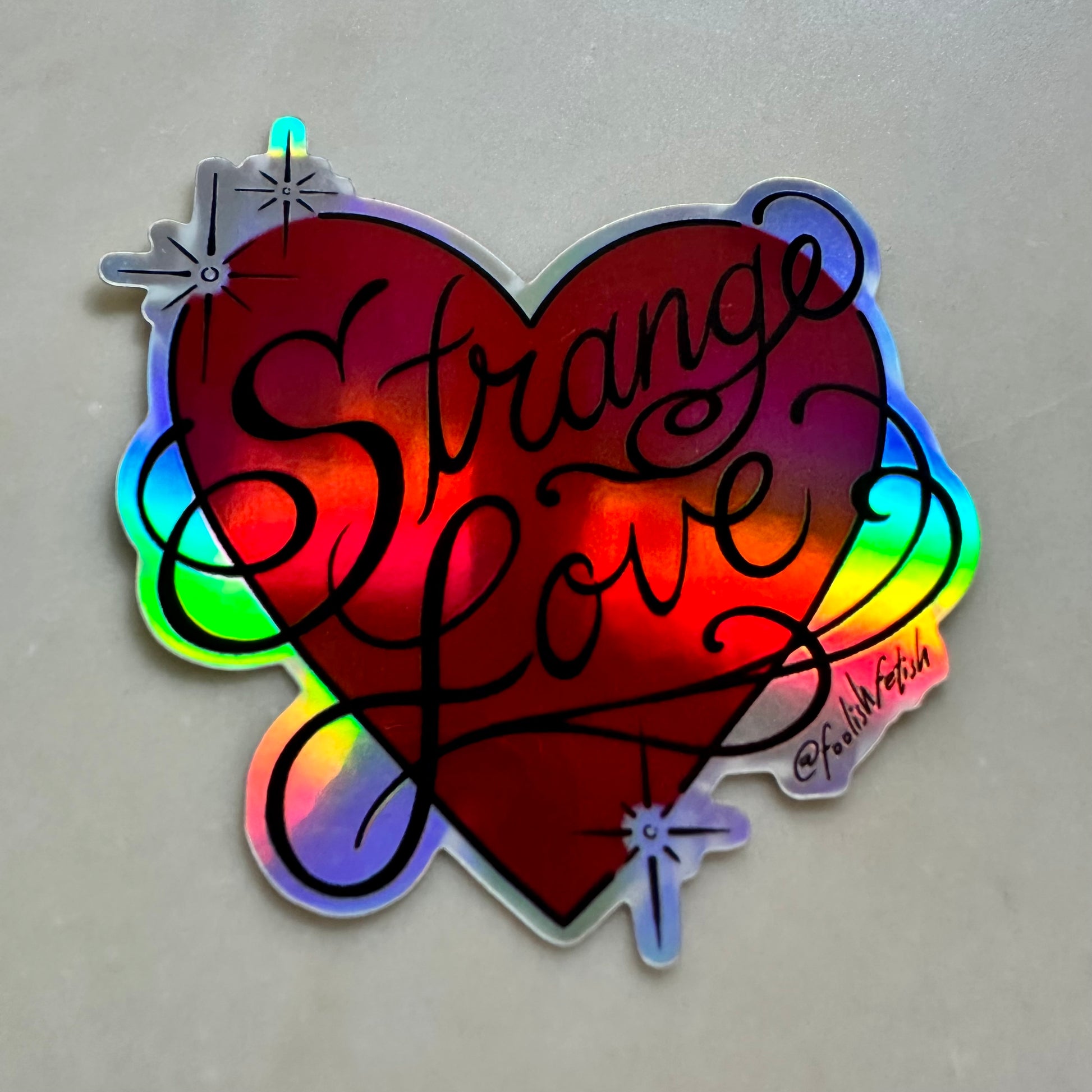 A holographic heart-shaped sticker with the words 'Strange Love' written in black script, photographed on a neutral gray surface.