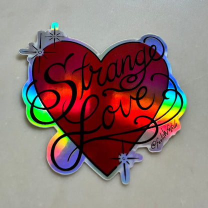A holographic heart-shaped sticker with the words 'Strange Love' written in black script, photographed on a neutral gray surface.