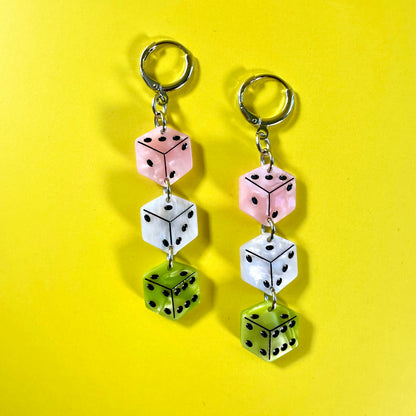 A pair of dangling earrings made from three stacked dice charms in pink, white, and green, attached to silver-tone rings, set against a solid yellow background.