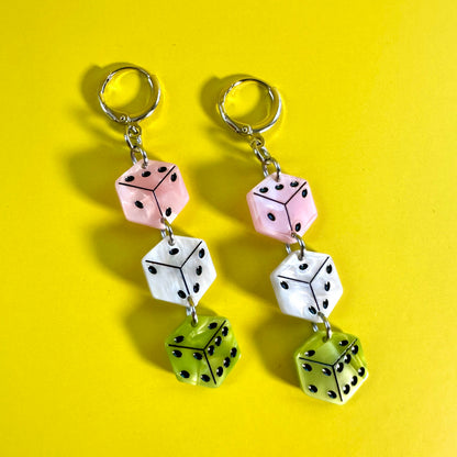 A pair of dangling earrings with three dice-shaped charms in pink, white, and green, attached to silver key rings, displayed on a bright yellow background.