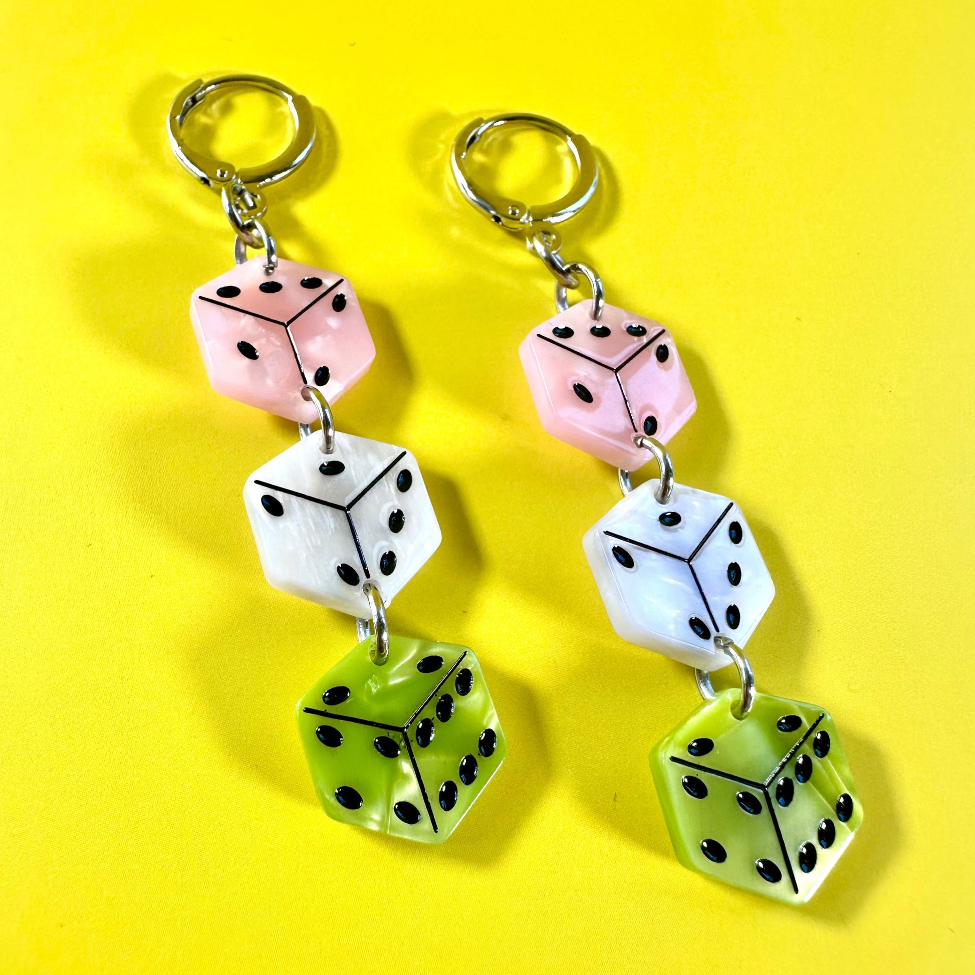 A pair of dangling earrings made from three stacked dice charms in pink, white, and lime green, attached to silver-tone split rings, displayed on a bright yellow background.