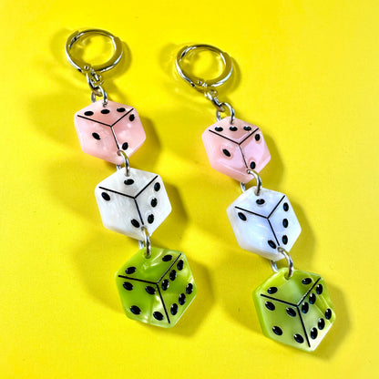 A pair of dangling earrings made from three stacked dice charms in pink, white, and lime green, attached to silver-tone split rings, displayed on a bright yellow background.