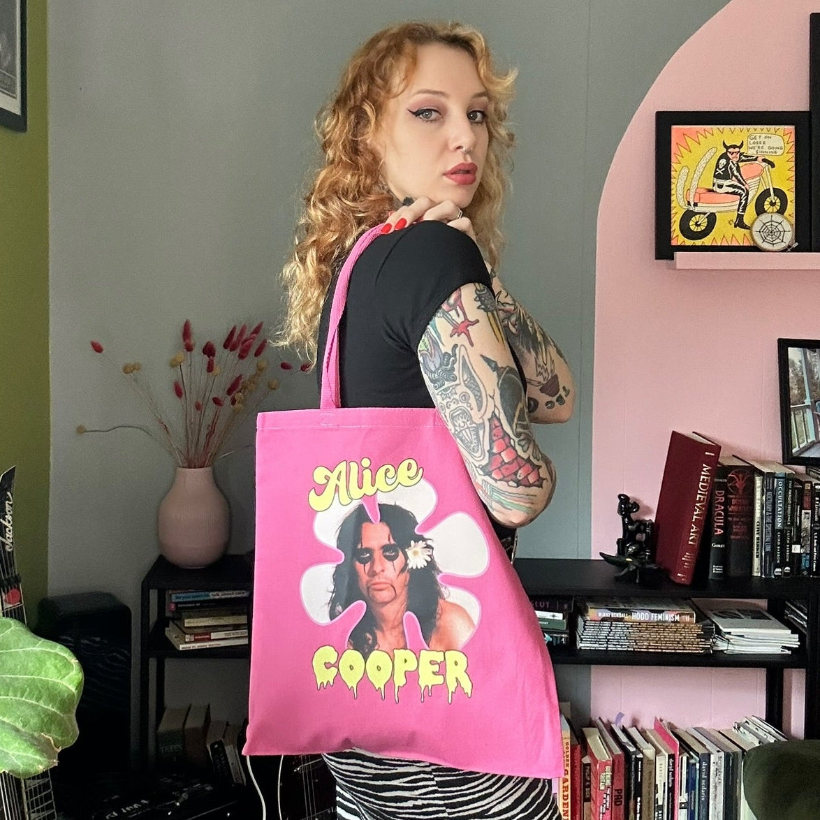 Person holding a pink tote bag with a graphic design in a room with green walls and a bookshelf.