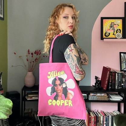 Person holding a pink tote bag with a graphic design in a room with green walls and a bookshelf.