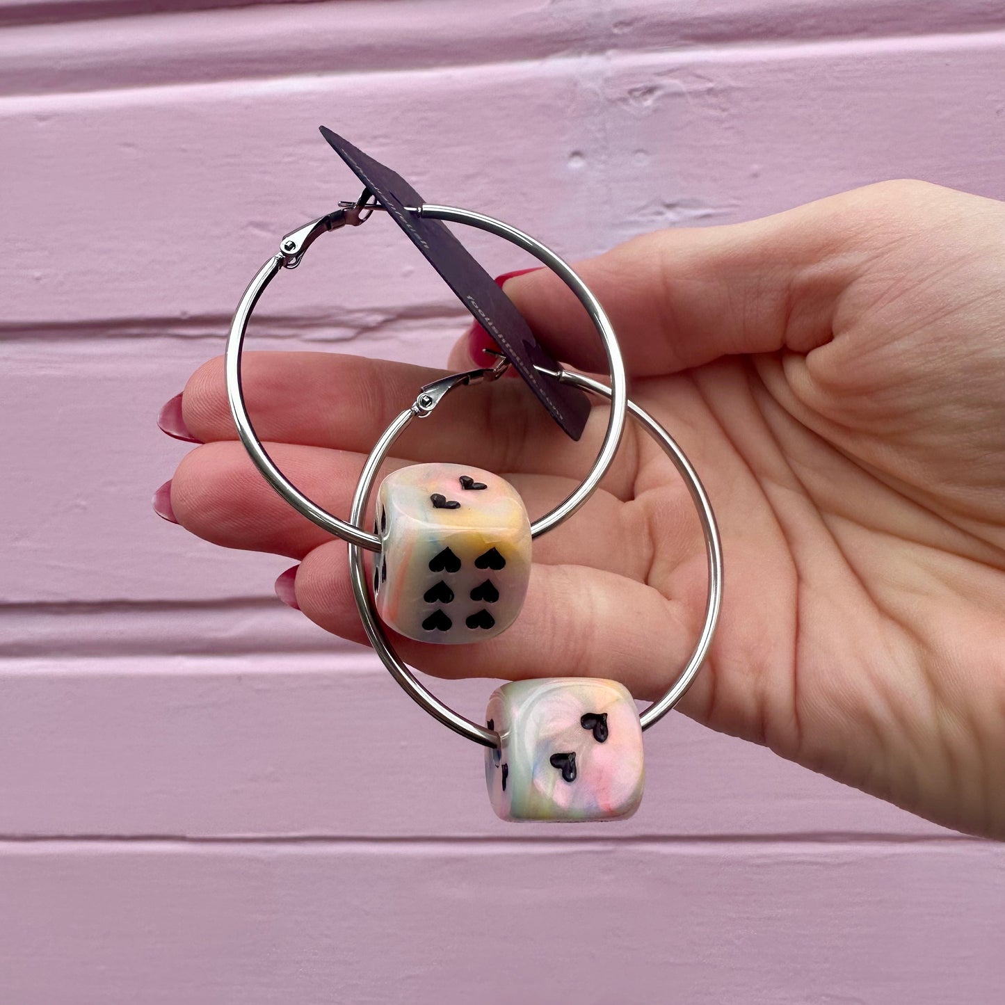 A pair of silver-colored hoop earrings with dice attached, displayed against a pink background