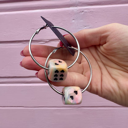 A pair of silver-colored hoop earrings with dice attached, displayed against a pink background