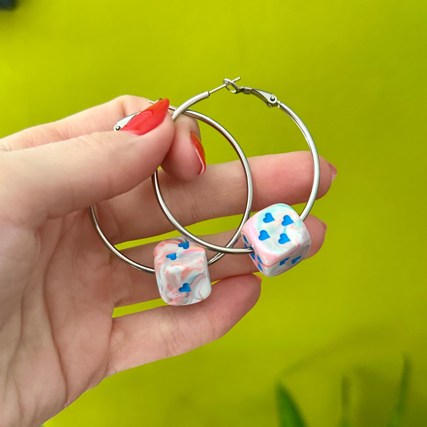 Hand holding silver hoop earrings with dice-shaped charms against a green background
