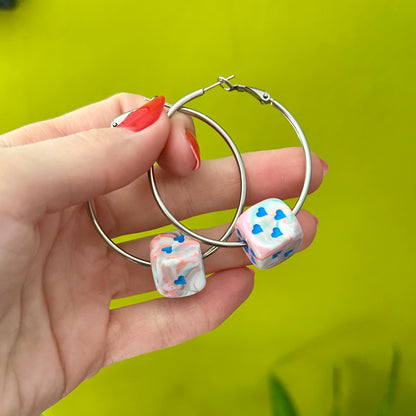 Hand holding silver hoop earrings with dice-shaped charms against a green background