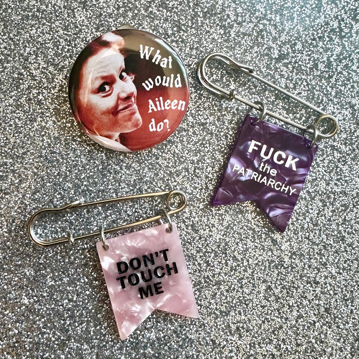 two safety pin banner pins in pink and purple that say Fuck the Patriarchy and Don't touch me. Plus a button pin of Aileen Wournos that says "What would Aileen do?"