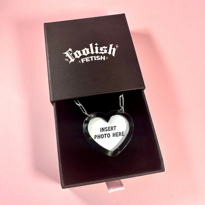 Heart-shaped photo insert pendant in a black box with 'Foolish Fetish' branding on a pink background.