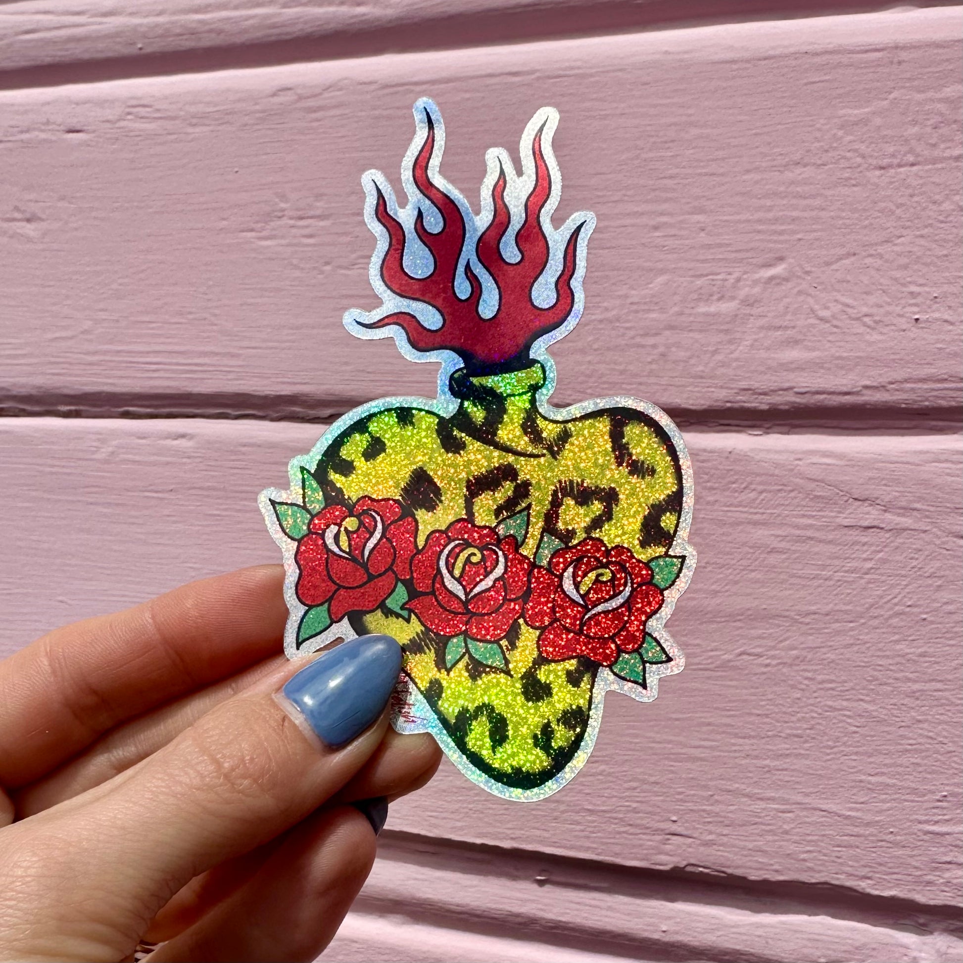 A hand with blue nail polish holds a die-cut sticker featuring a glittery yellow leopard-print heart with three red roses and red flames, set against a pink wooden background.