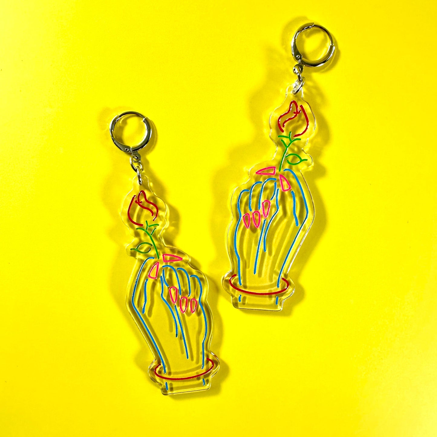 A pair of dangling acrylic earrings featuring a stylized hand holding a flower, shown on a bright yellow background.