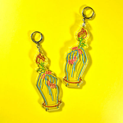 A pair of dangling acrylic earrings featuring a stylized hand holding a flower, shown on a bright yellow background.