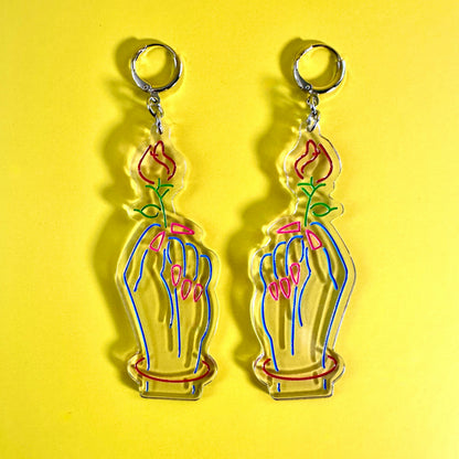 A pair of clear acrylic earrings charms shaped like hands holding pink flowers, shown on a bright yellow background.