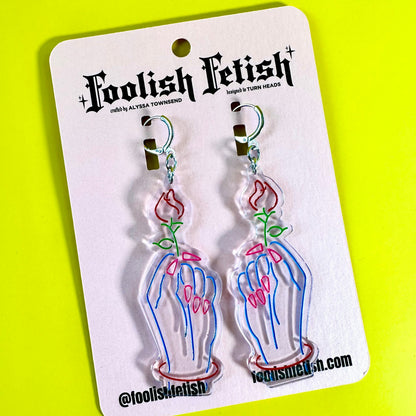 A pair of dangling earrings on a white retail card. The earrings are made of clear acrylic with a printed neon-style design of skeletal hands holding pink roses with green stems.