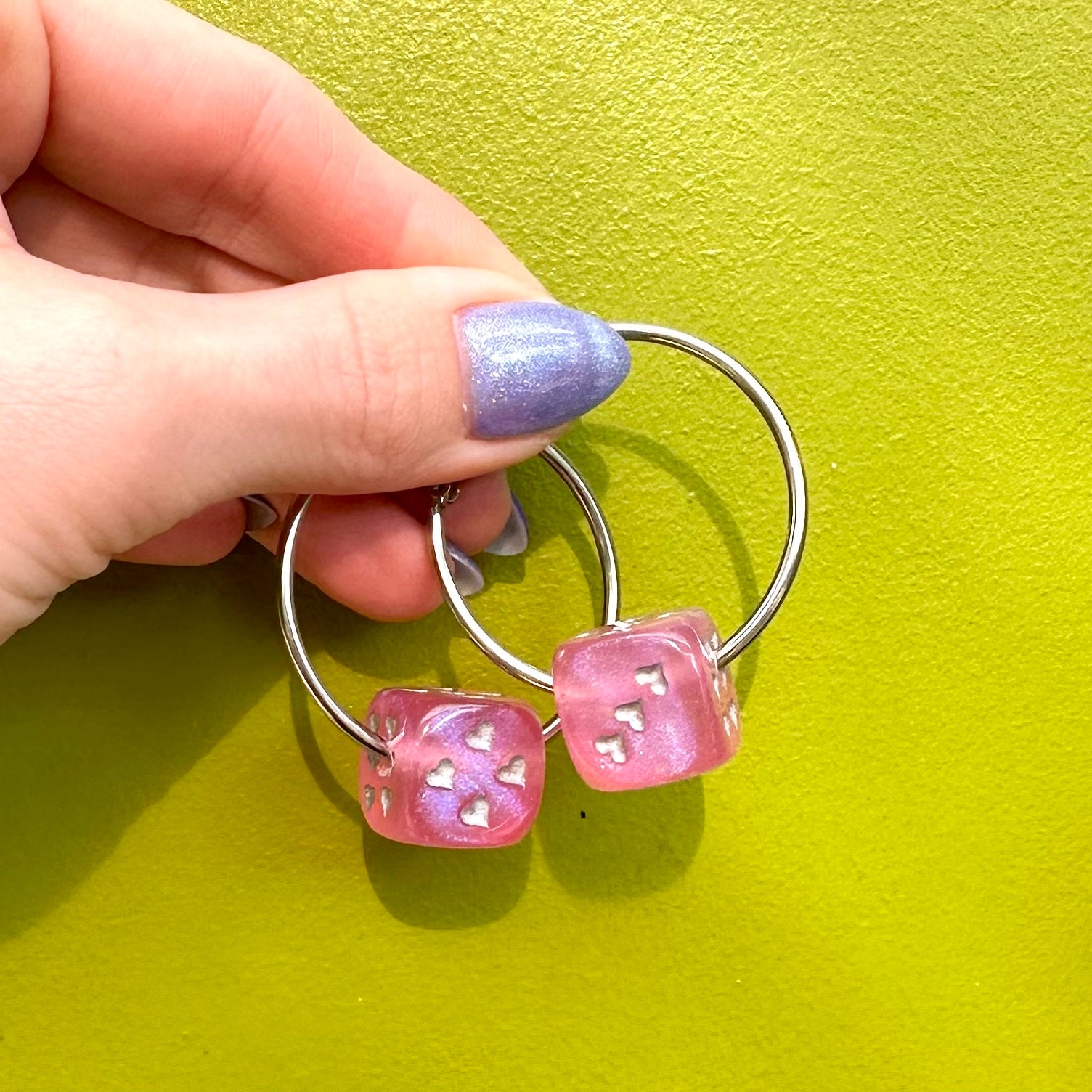 A pair of silver-colored hoop earrings with dice attached, displayed against a pink background