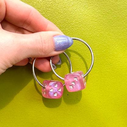 A pair of silver-colored hoop earrings with dice attached, displayed against a pink background