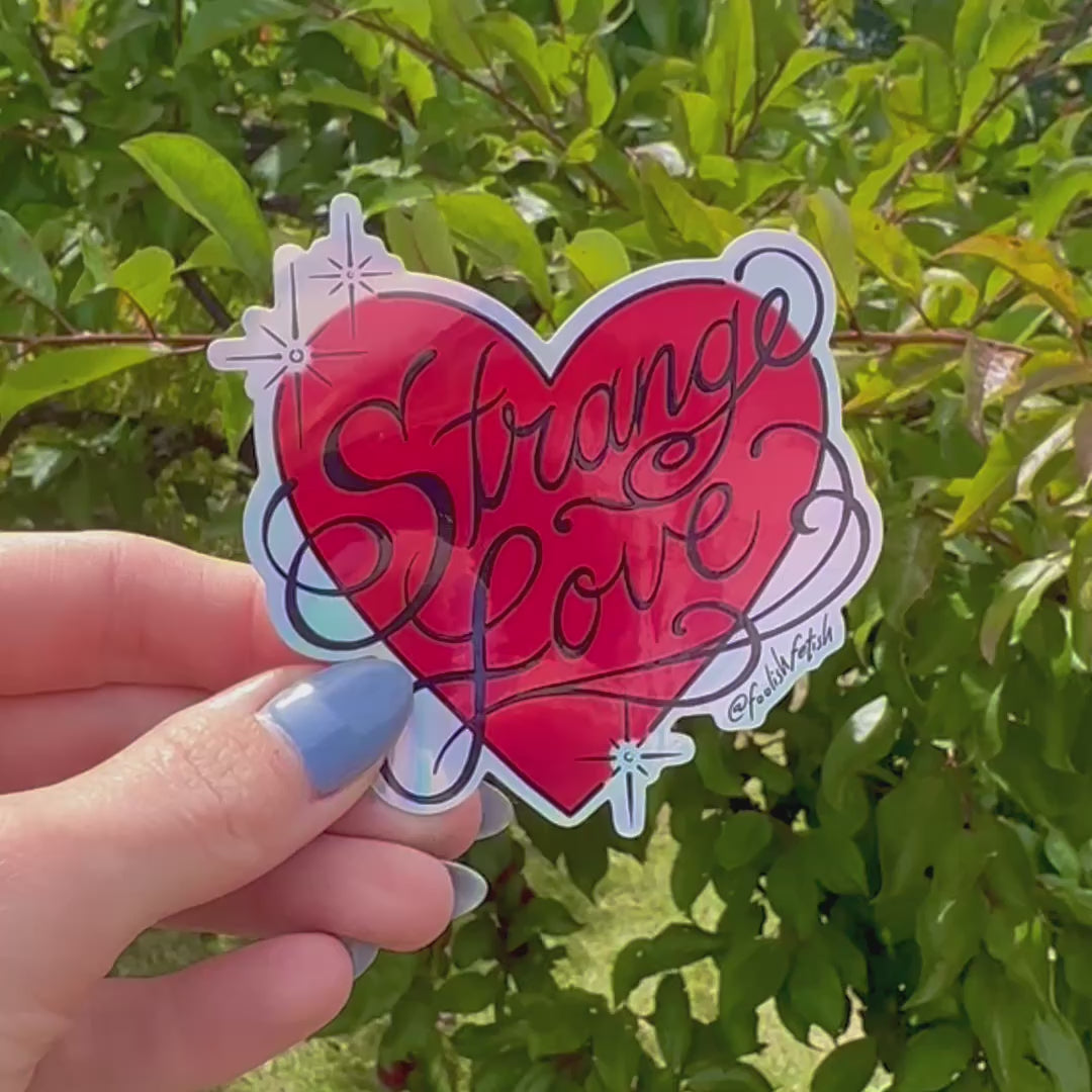 Strange Love Heart Sticker video of hand holding it outside against green plants background
