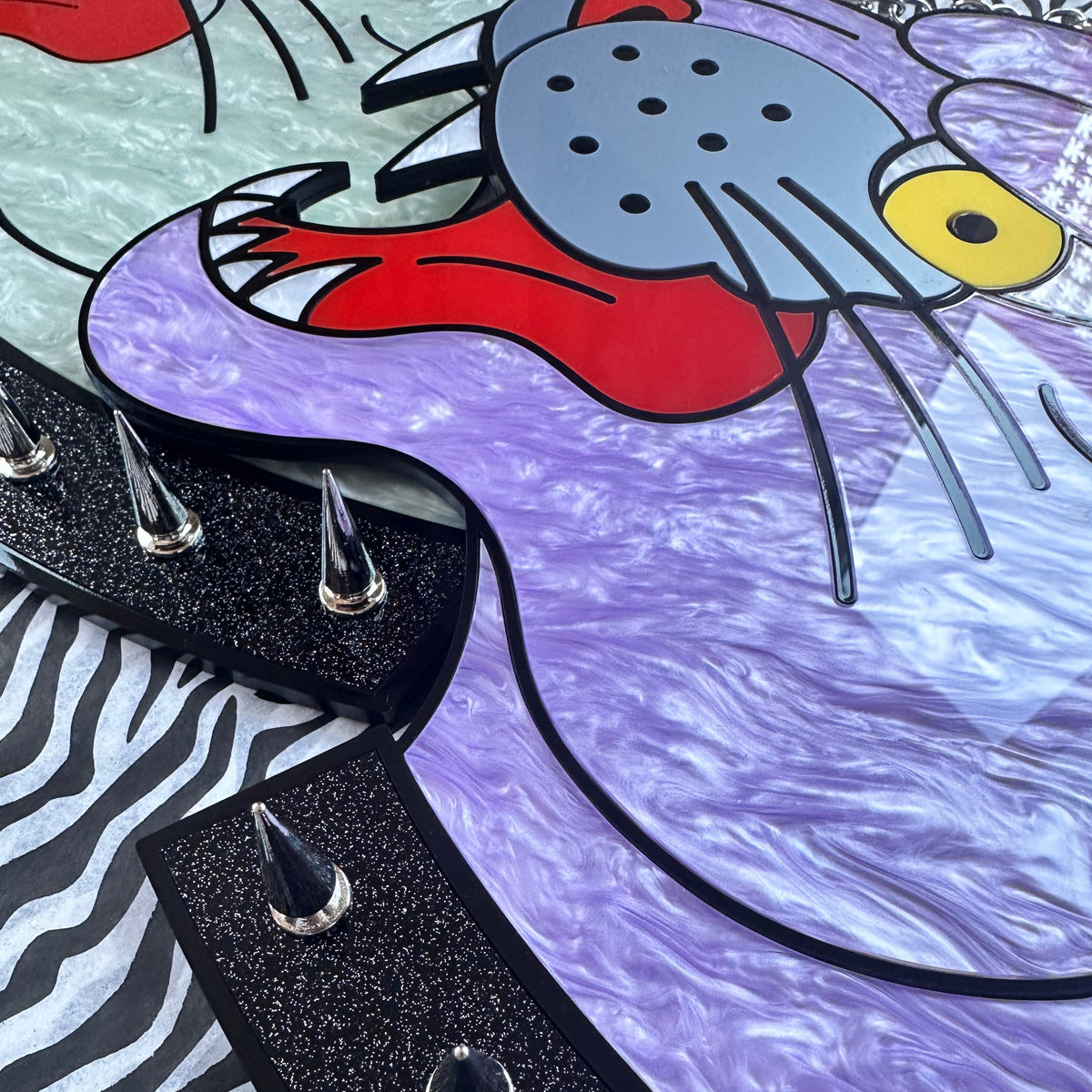 Close-up of a colorful graphic surface featuring a stylized cat face in gray and red, a purple marbled curved shape, black spiked trim, a black-and-white zebra pattern, and a light blue background.