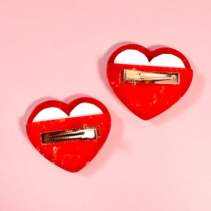Two red heart-shaped hair clips with gold bars on a pink background