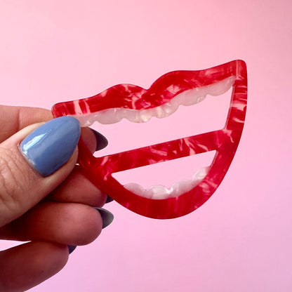 A hand with blue nail polish holds a red and white marbled lip-shaped scarf ring against a solid pink background.