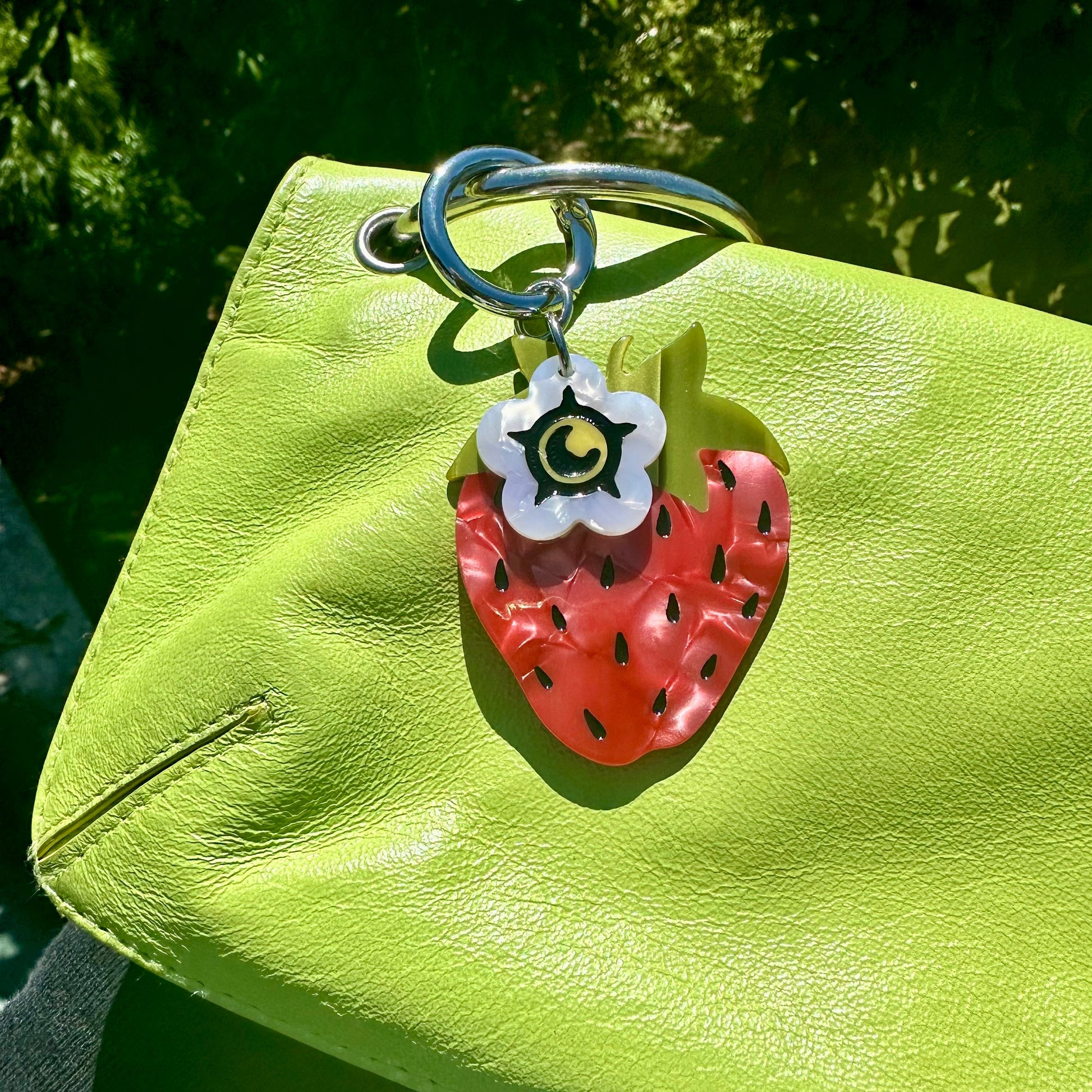 red strawberry bag charm with small white flower blossom charm on green purse with trees in background