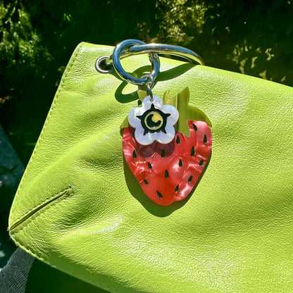 red strawberry bag charm with small white flower blossom charm on green purse with trees in background