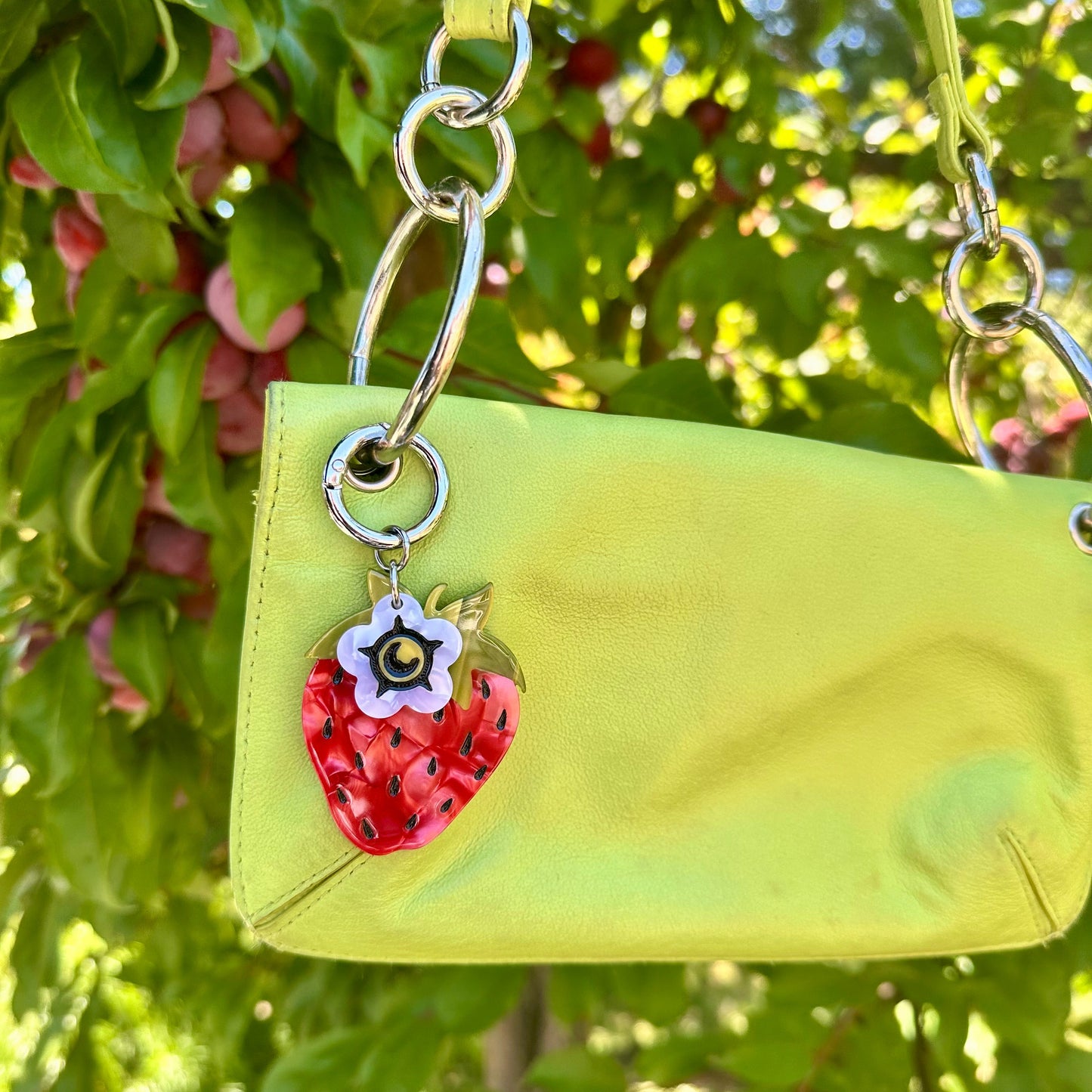 red strawberry bag charm with small white flower blossom charm on green handbag with plants in the background