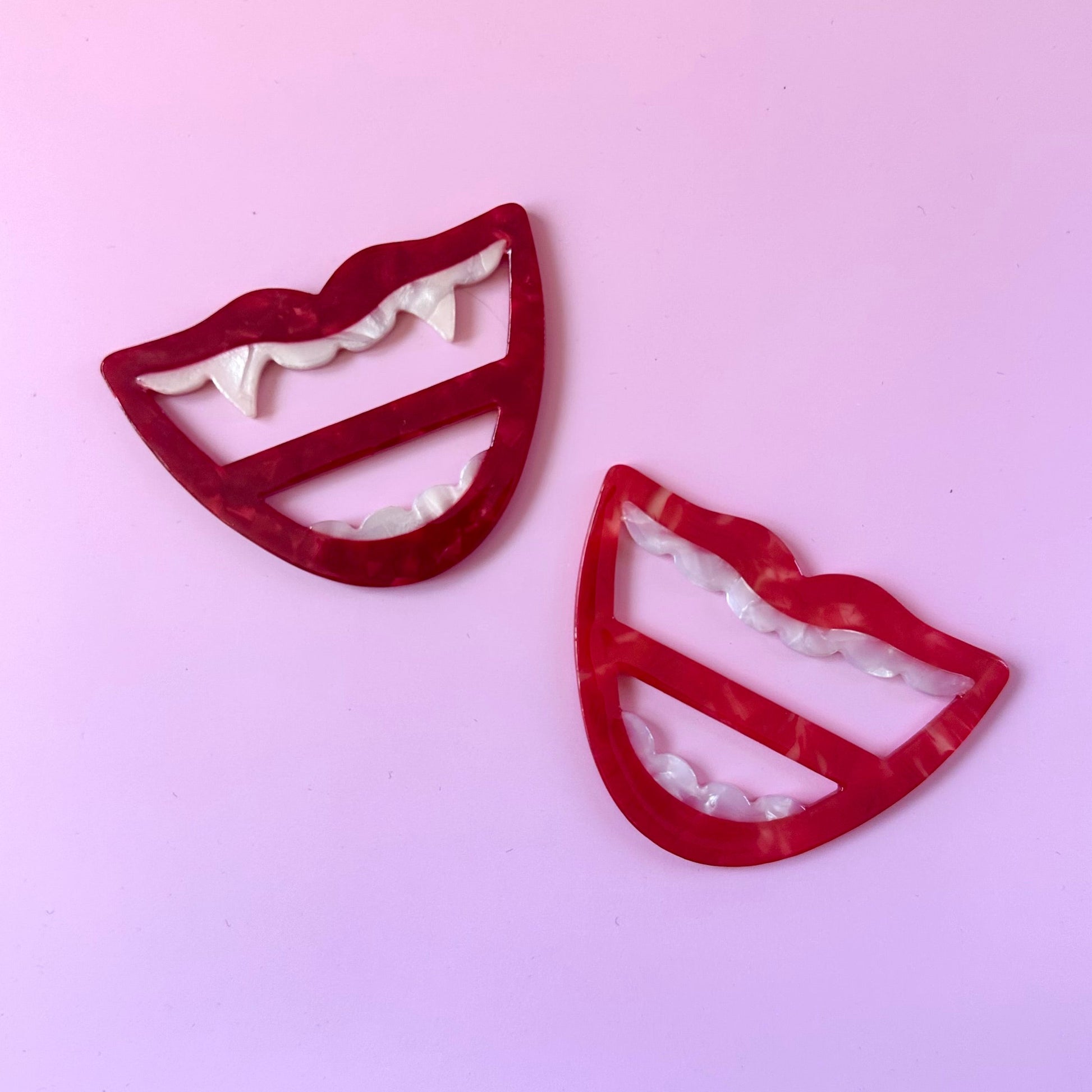 Two red lips-shaped scarf rings with white teeth details, displayed on a solid light pink background.
