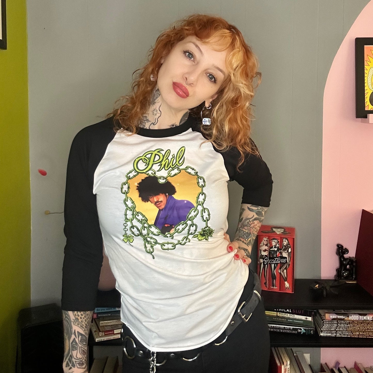 Person wearing a white and black baseball shirt with a graphic design, standing in a room with books and a pink wall.