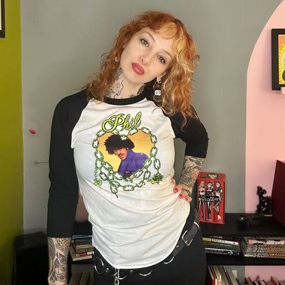 Person wearing a white and black baseball shirt with a graphic design, standing in a room with books and a pink wall.