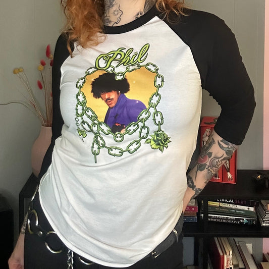 Person wearing a white and black baseball shirt with a graphic design, standing in a room with a pink wall and books.